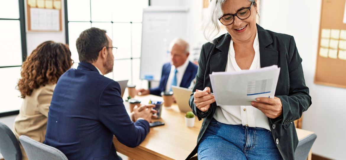 Group of middle age business workers working at the office. Woman smiling happy holding documents sitting on the table.