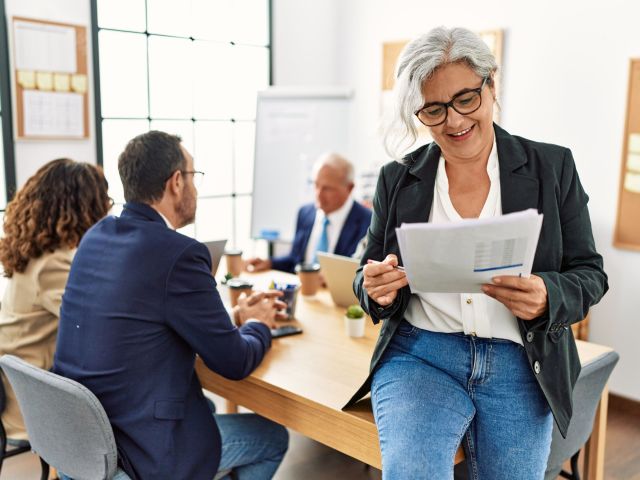 Group of middle age business workers working at the office. Woman smiling happy holding documents sitting on the table.