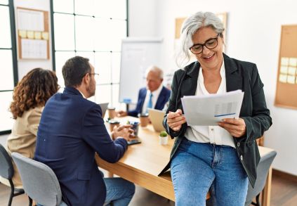 Group of middle age business workers working at the office. Woman smiling happy holding documents sitting on the table.